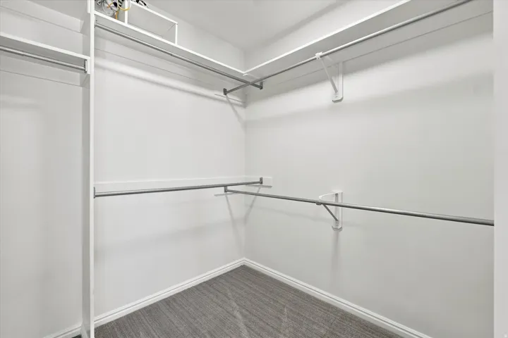 Walk-in closet featuring white painted walls, white shelving, chrome hanging rods, and grey carpet flooring