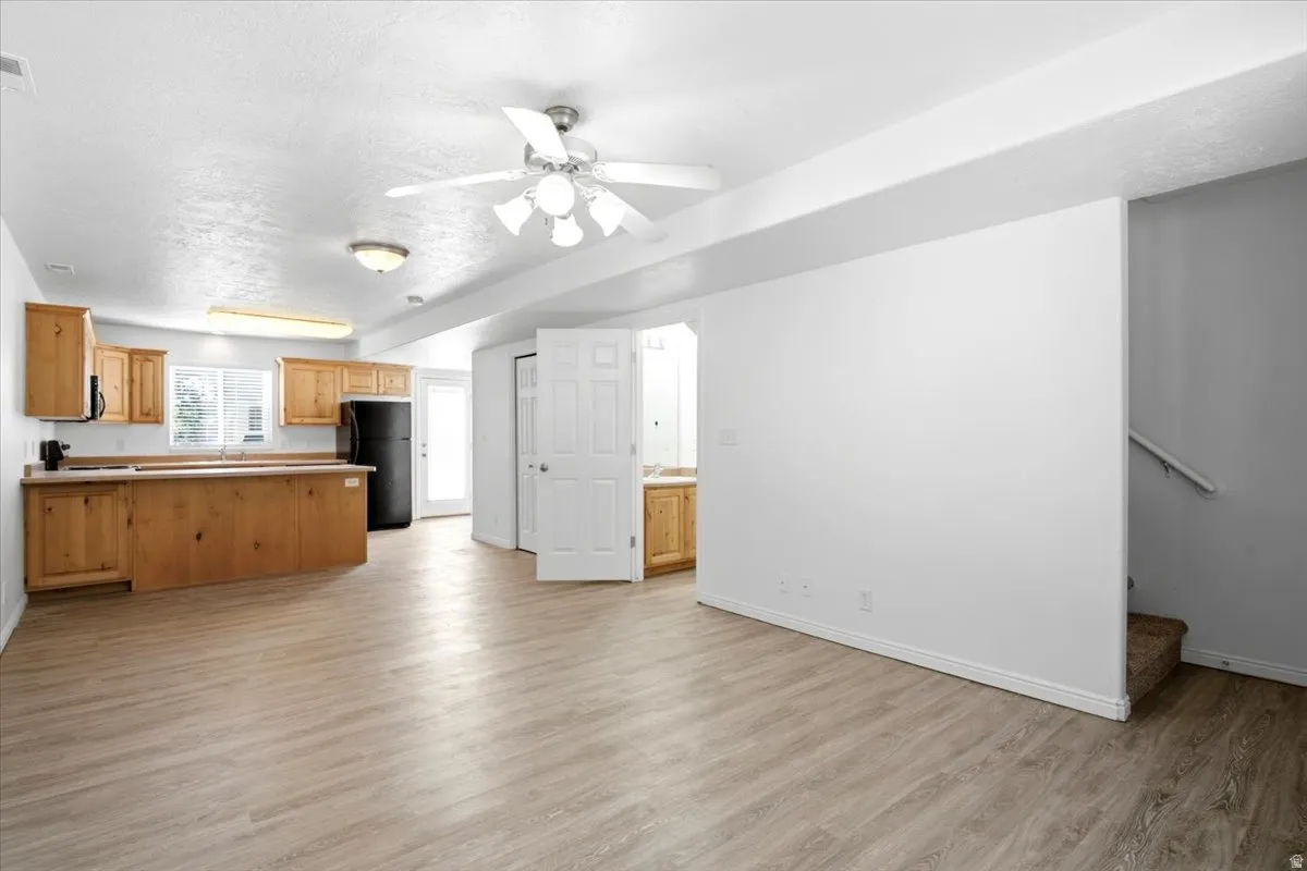 Open-concept living area featuring wood-finish flooring, a kitchen with light wood cabinetry and a black refrigerator, and a ceiling fan with integrated lighting