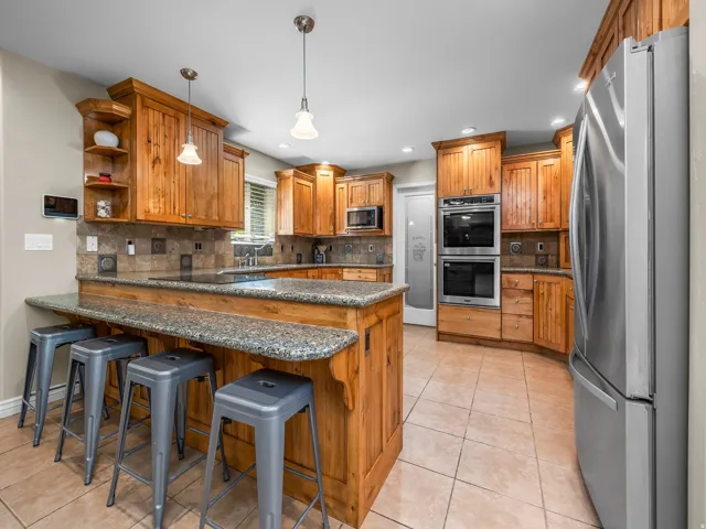 Kitchen featuring warm wood cabinetry, a granite-finish breakfast bar, and stainless steel appliances including double ovens and a full-size refrigerator