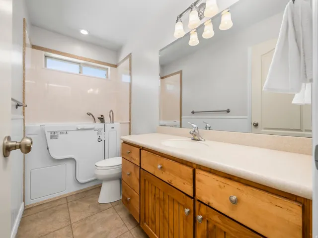 Bathroom featuring a walk-in tub, wood-finish vanity with an integrated sink, large mirror, and tile flooring