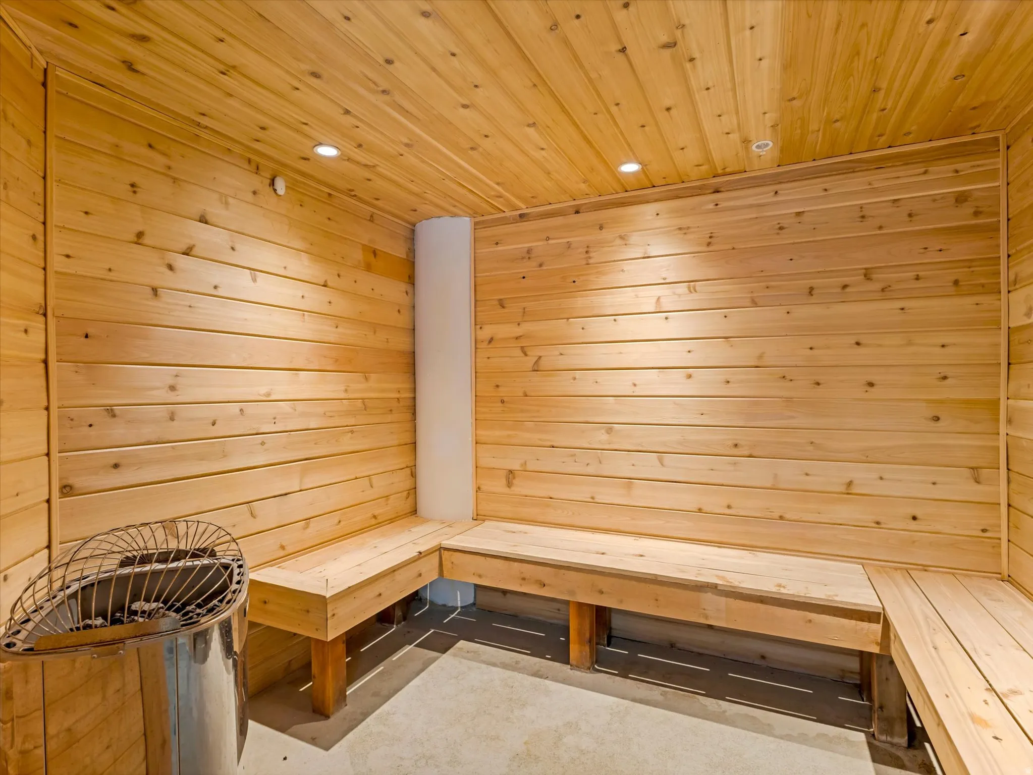Wood-paneled sauna featuring built-in benches, recessed lighting, concrete flooring, and a stainless steel heater