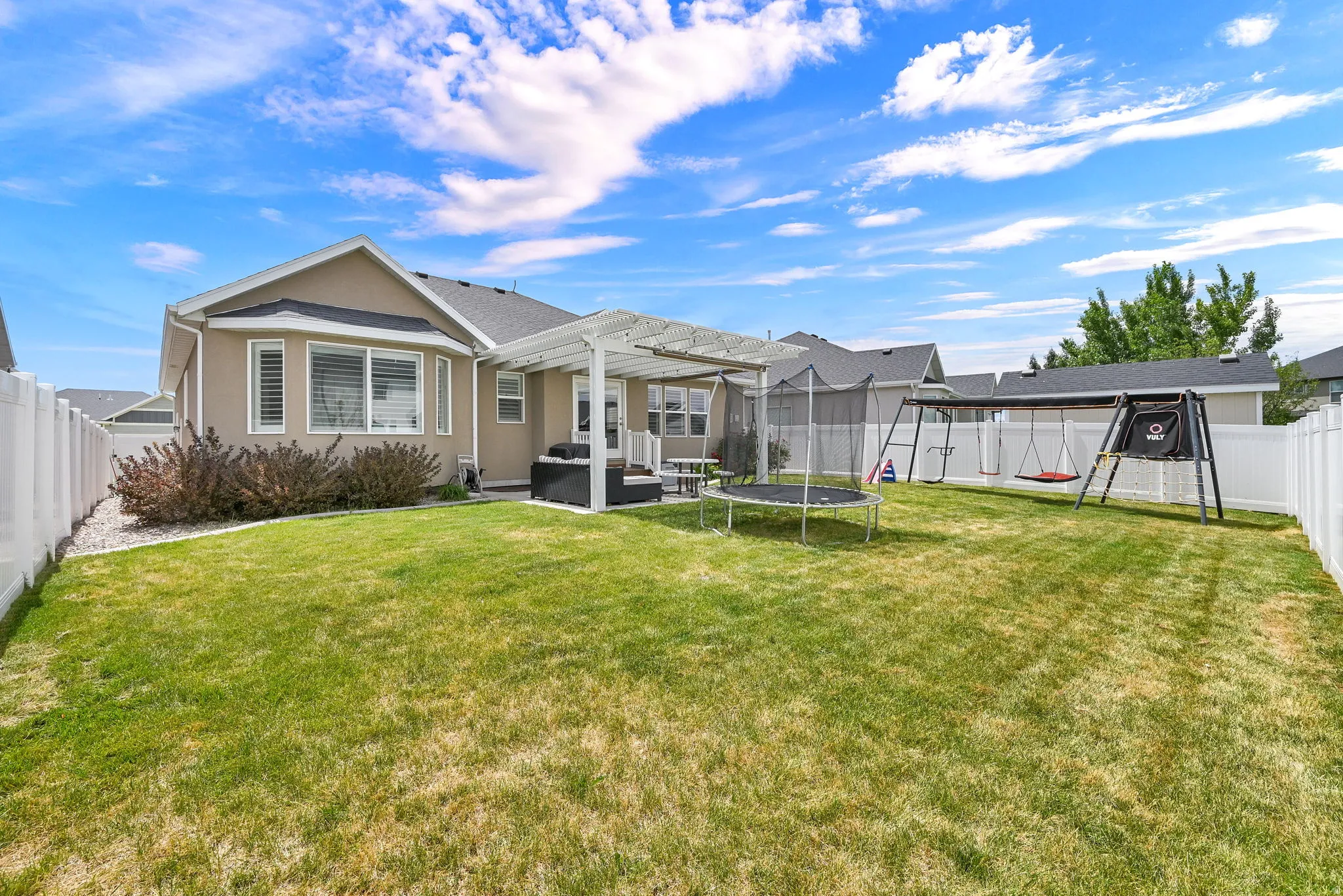 Expansive backyard featuring a pergola-covered patio, mature shrubbery, a manicured lawn, and white vinyl fencing