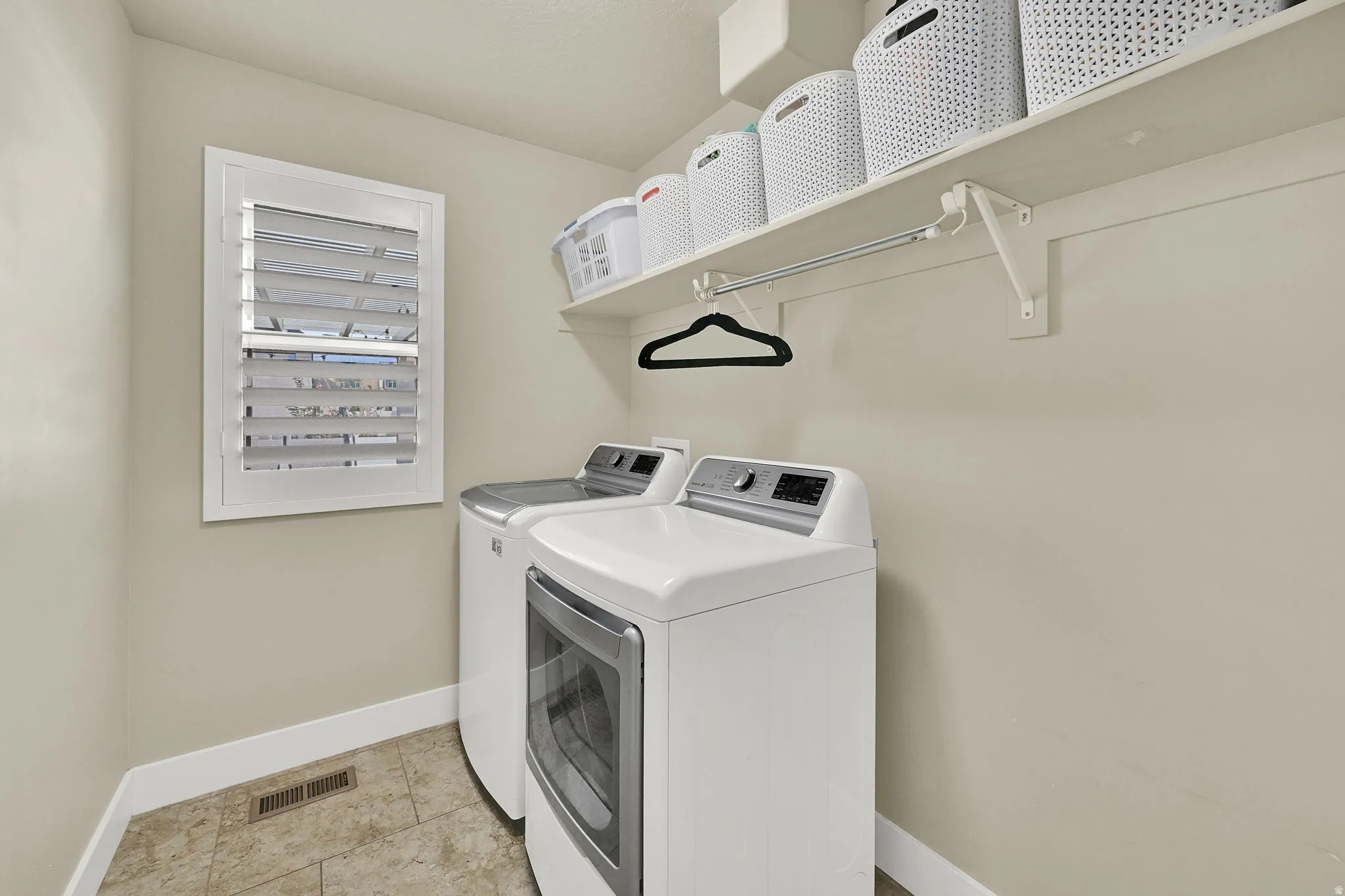 Dedicated laundry room featuring tile flooring, a window with white louvered shutters, and a built-in shelving system with hanging rod