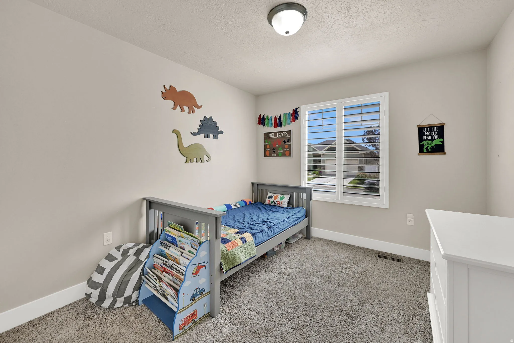 Light-filled room featuring a broad window with white plantation shutters, neutral wall tones, textured carpeting, a flush-mount ceiling light, and white baseboards