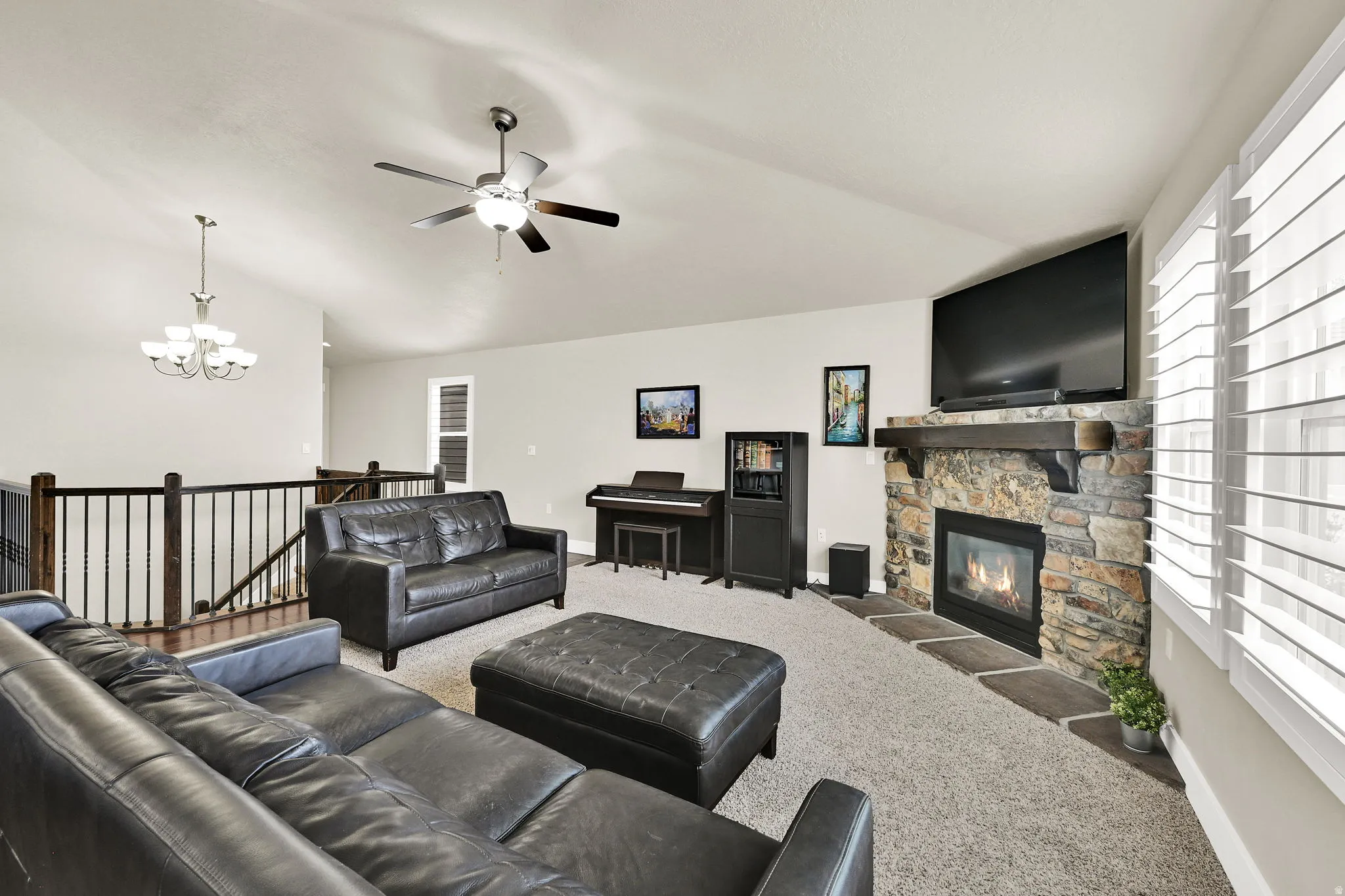 Vaulted ceiling living room featuring a stone-faced fireplace with a wood mantel, a ceiling fan with integrated lighting, and plantation shutters