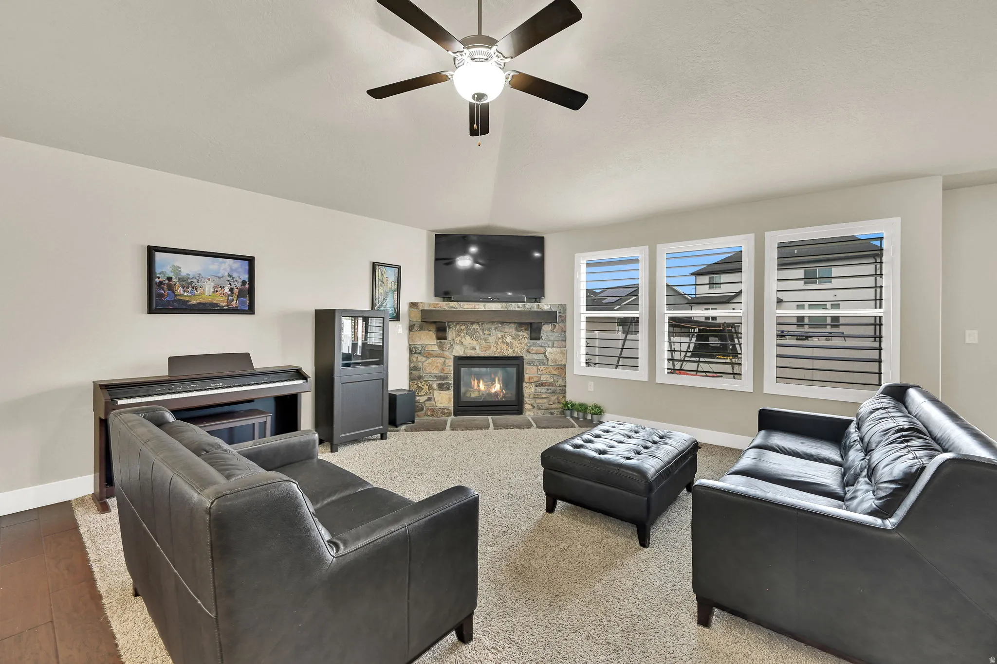 Living area featuring a stone-faced fireplace with a wood mantel, a ceiling fan with integrated lighting, and three large windows