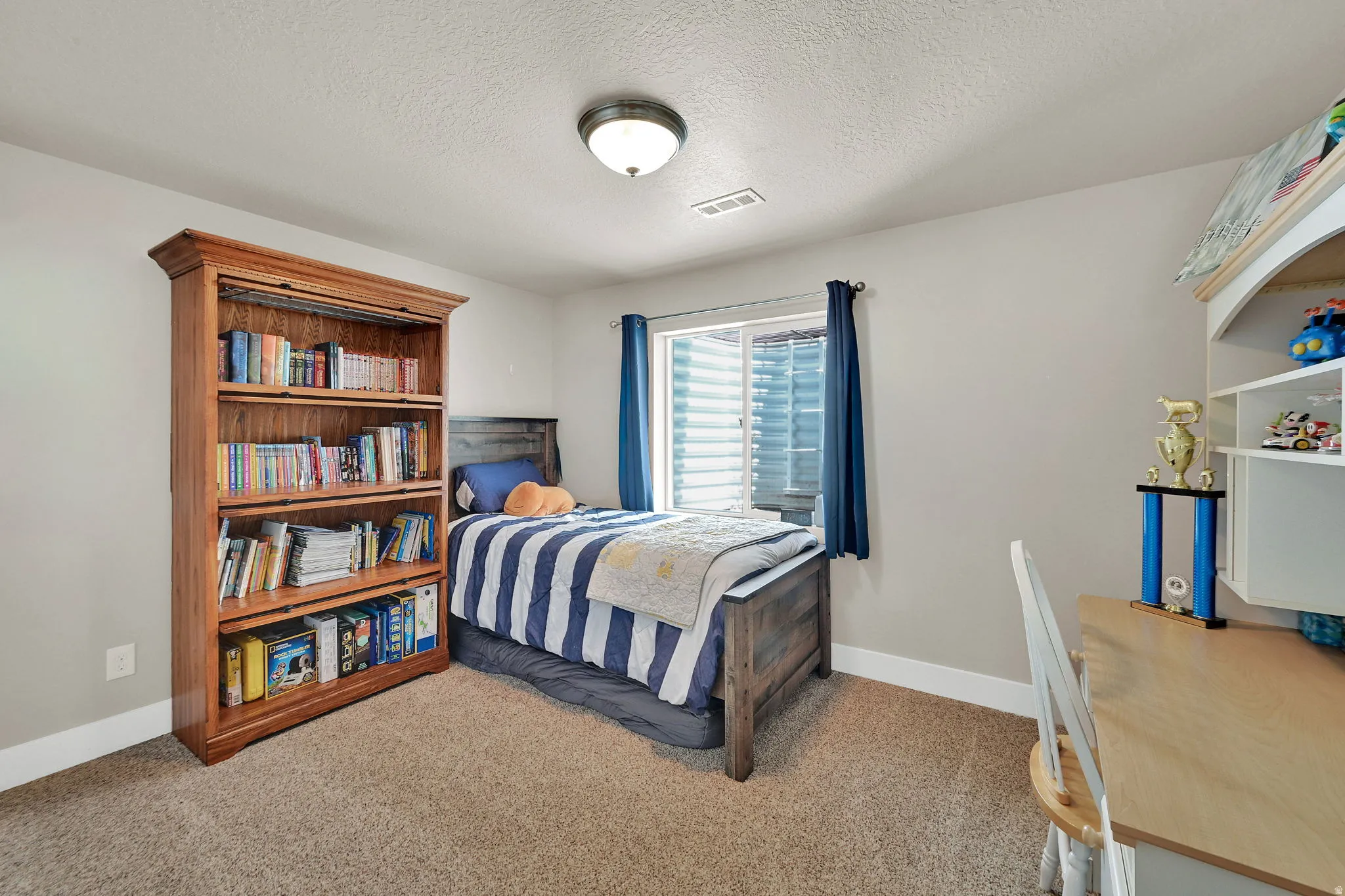 Bedroom featuring light neutral wall paint, a single window with blinds and drapes, and textured carpet flooring