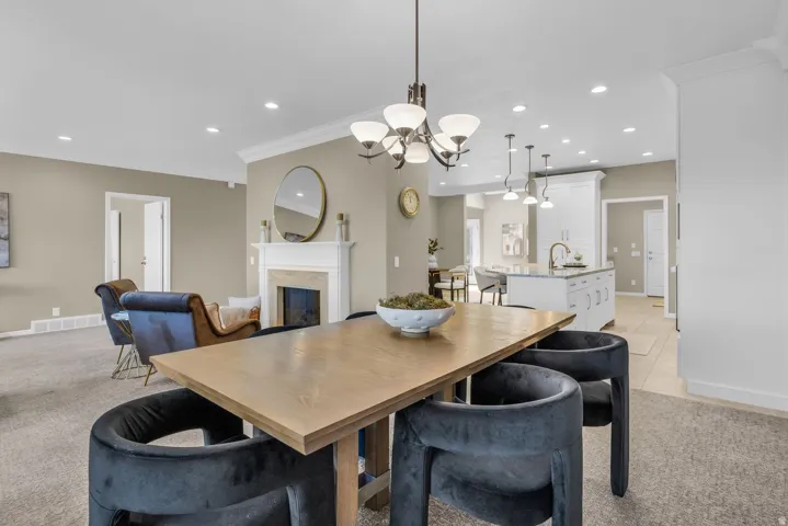 Open-concept living space featuring a dining area with a wood-finish table, an adjacent kitchen with a central island and white cabinetry, a fireplace with a white mantel, recessed lighting, and crown molding