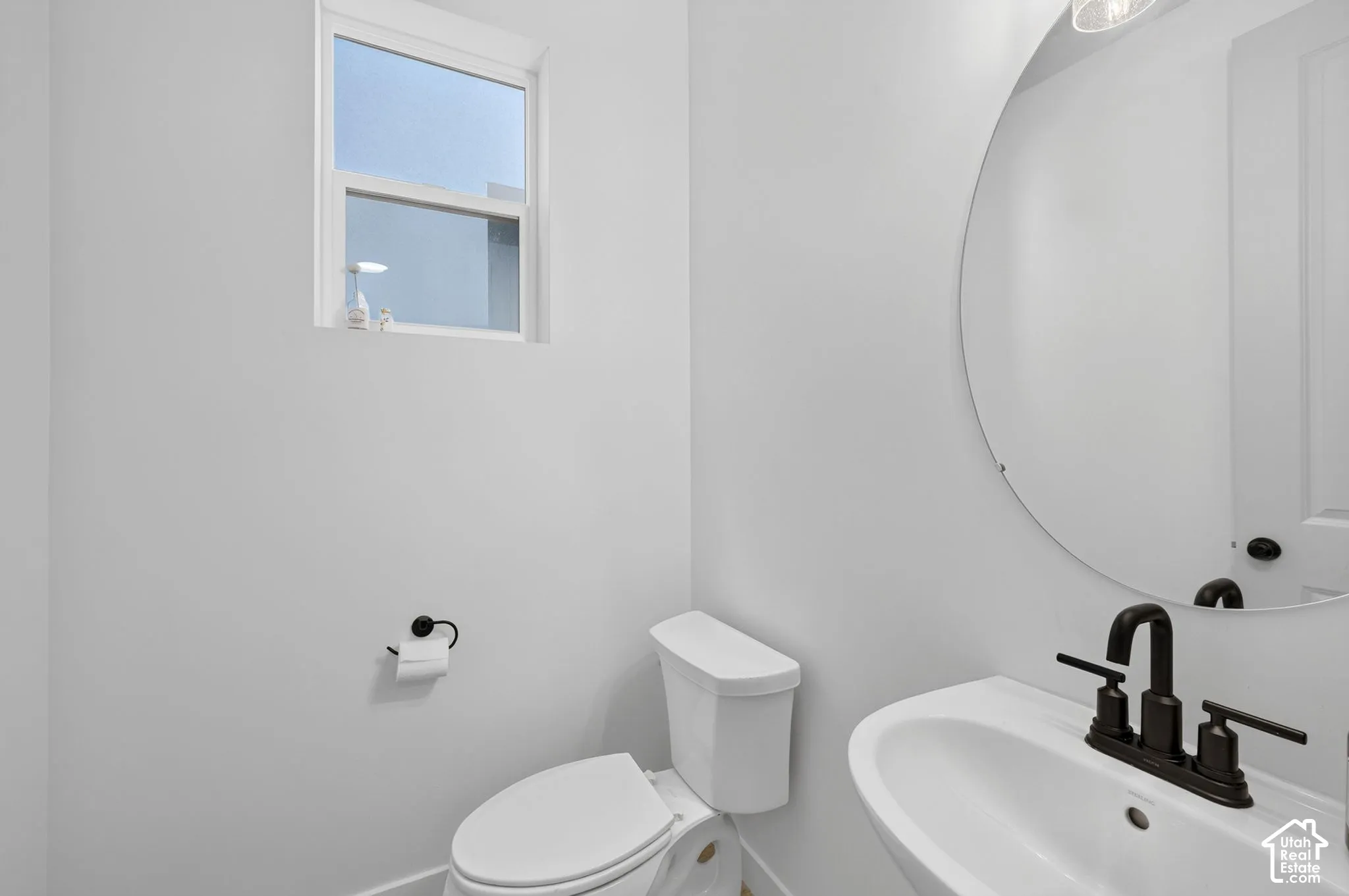 White-walled powder room featuring a pedestal sink with a matte black faucet, a round wall-mounted mirror, and a window for natural light