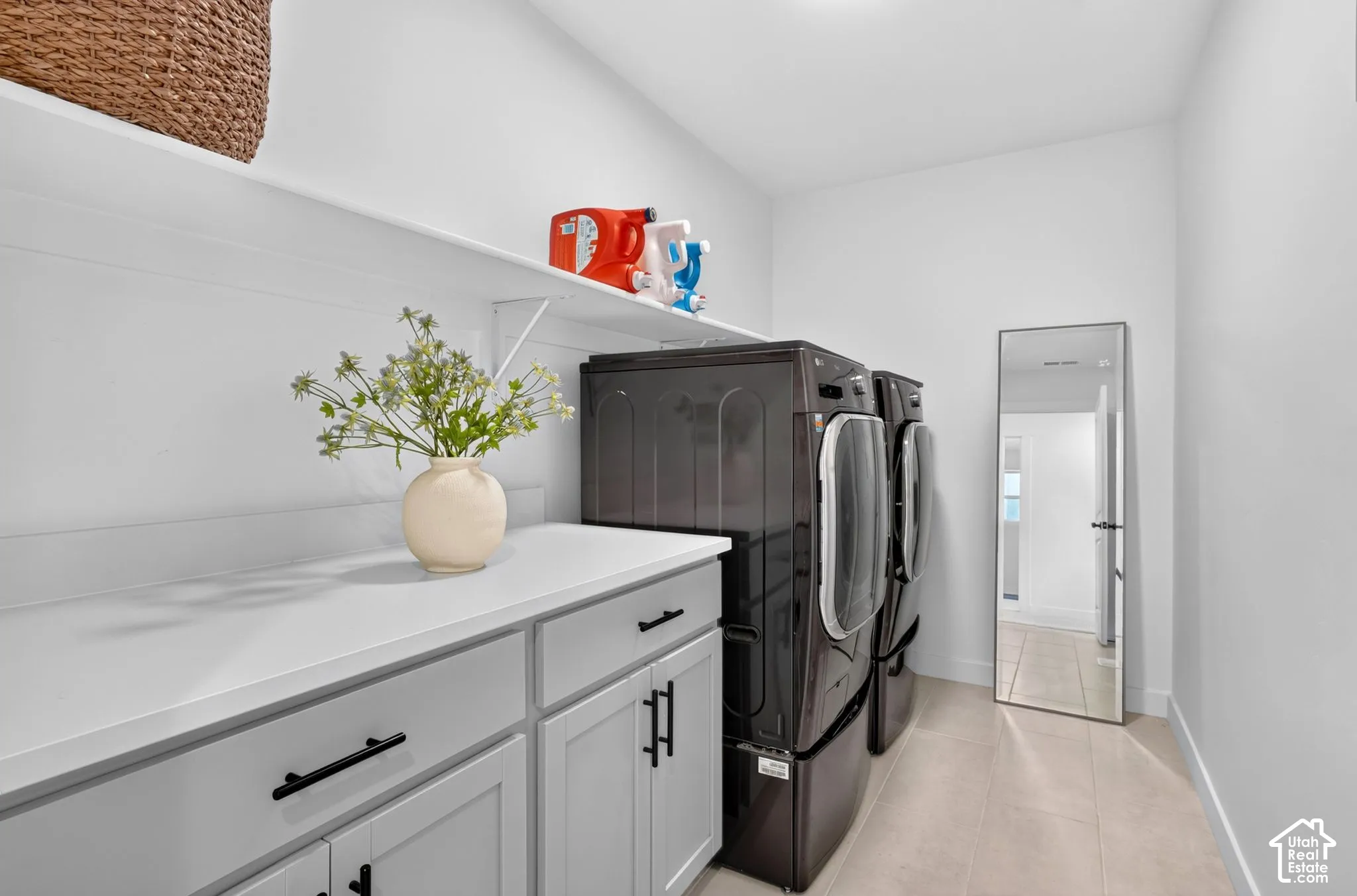 Dedicated laundry area featuring a light-toned countertop, light gray cabinetry with matte black hardware, and an overhead shelf
