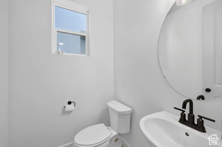 White-walled powder room featuring a pedestal sink with a matte black faucet, a round wall-mounted mirror, and a window for natural light