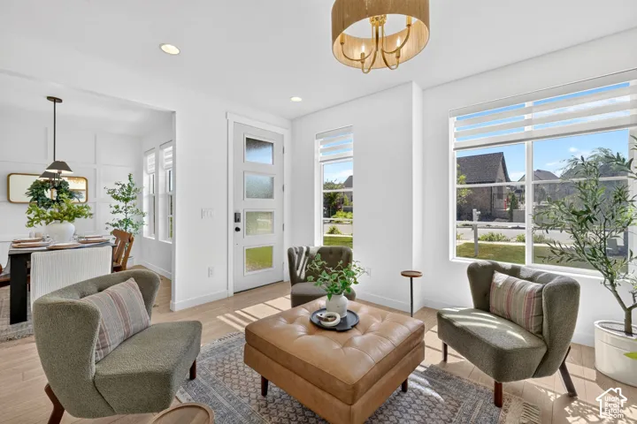 Bright interior space featuring wood-finish flooring, a contemporary chandelier, large windows with horizontal blinds, a modern entry door with frosted glass panels, and recessed lighting