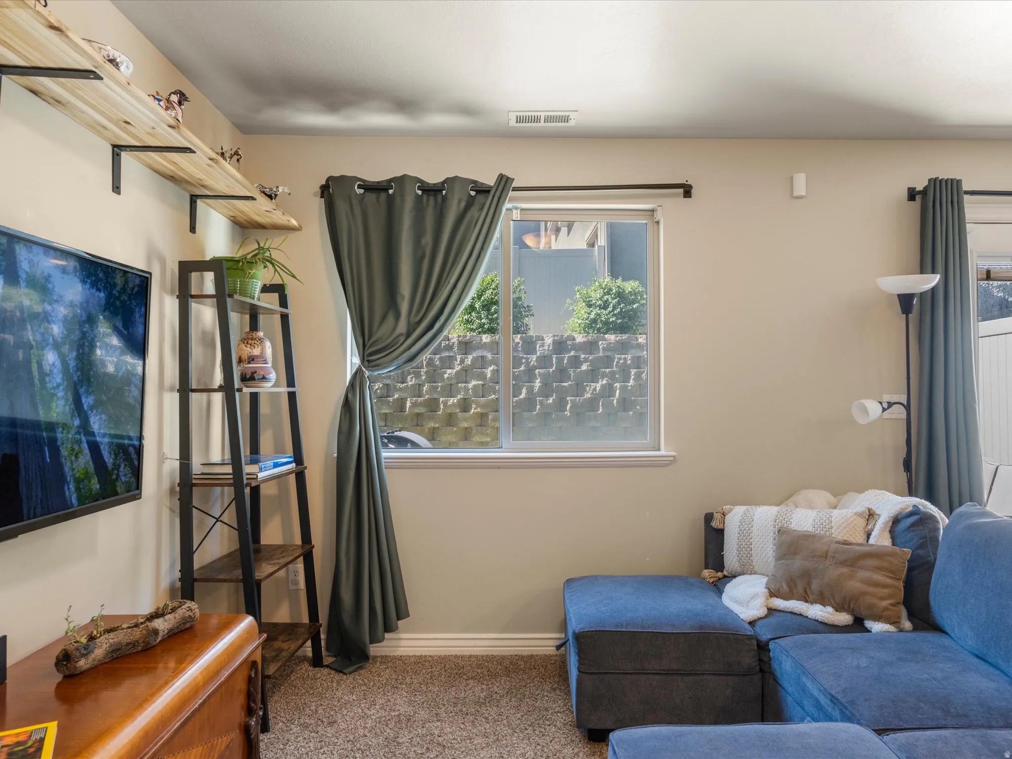 Warm interior room featuring neutral wall tones, textured carpeting, and a window with a stone retaining wall backdrop