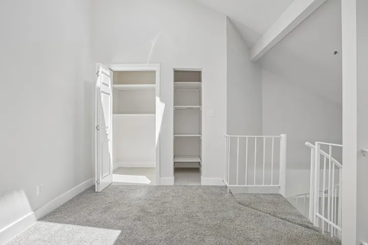 Carpeted loft area featuring a white metal railing, built-in shelving, a recessed closet with an open door, vaulted ceilings, and white painted walls