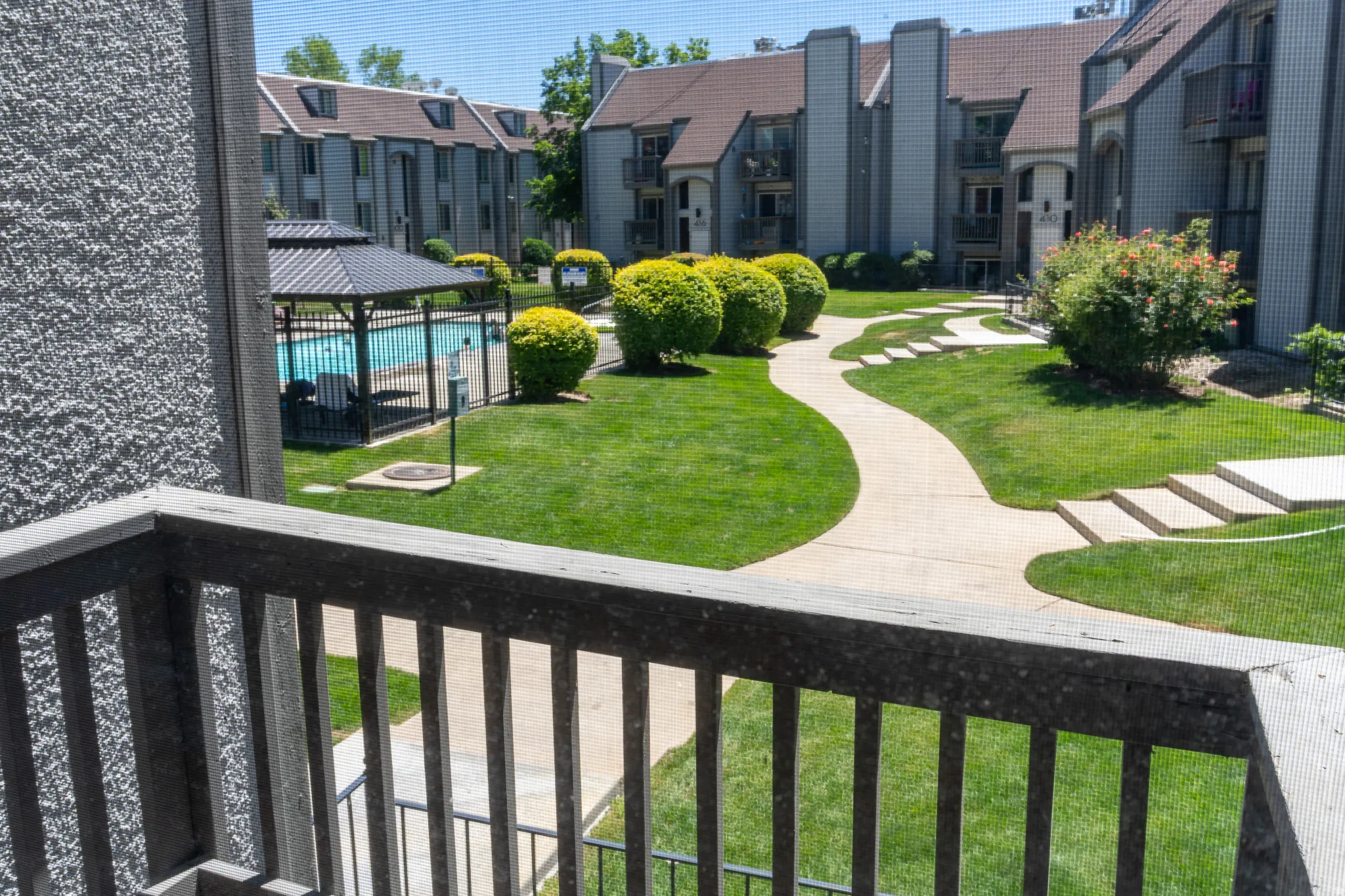 Exterior balcony overlooking a central courtyard with a swimming pool, covered gazebo, concrete pathways, and manicured green spaces