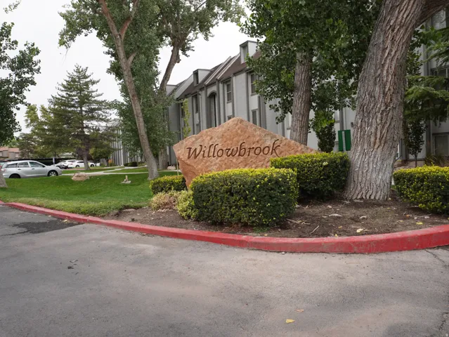Landscaped entry featuring a natural stone monument with engraved lettering, mature trees, manicured shrubs, and a verdant lawn