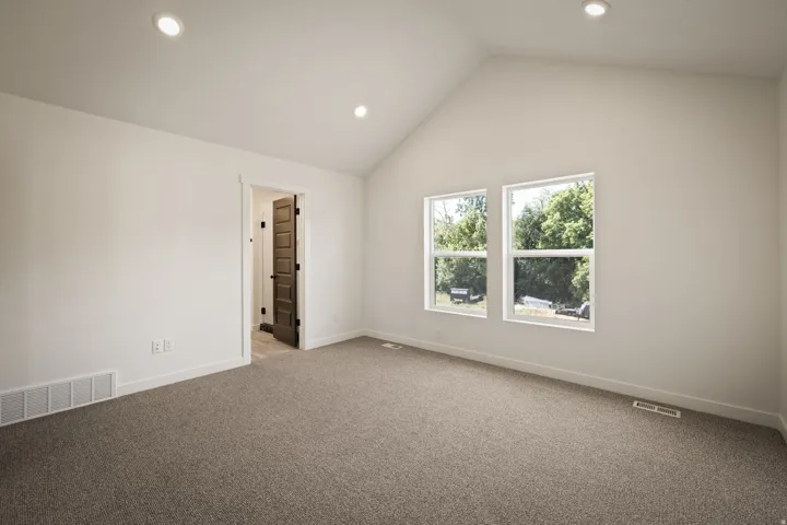 Spacious room featuring a vaulted ceiling with recessed lighting, two windows, wall-to-wall carpeting, and a dark paneled interior door