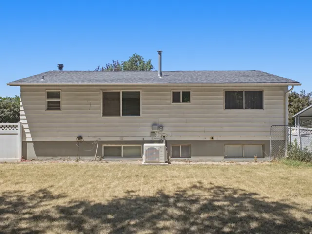 Rear exterior with horizontal siding, a dark shingled roof, multiple windows, a white lattice fence segment, and a chain-link fence
