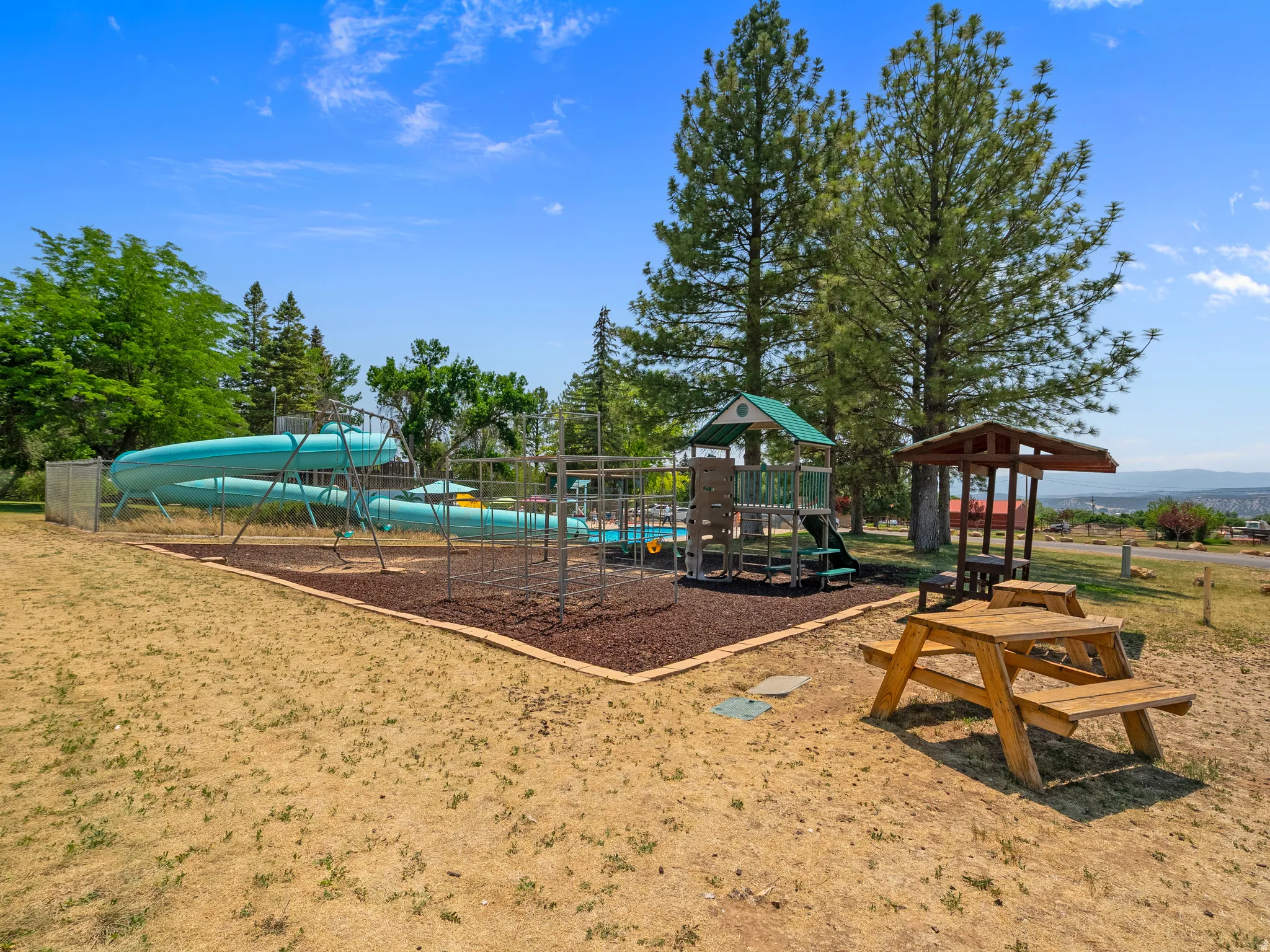 Outdoor recreational area featuring a large blue water slide, a structured play system with a green roof, a shaded picnic table, and additional wooden seating