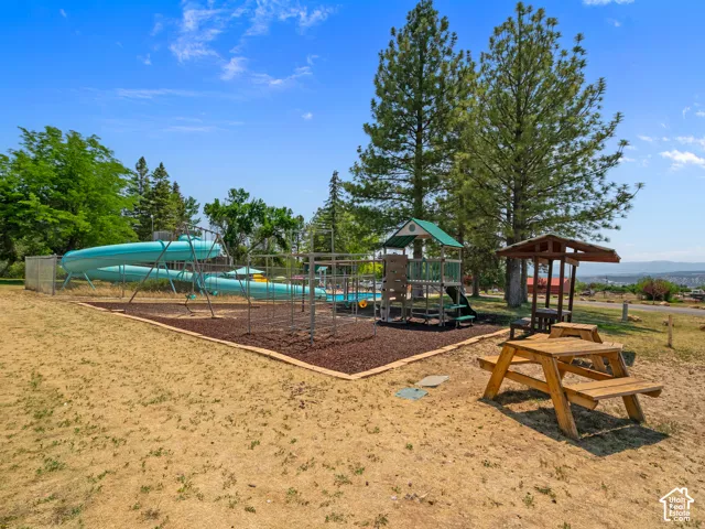 Outdoor recreational area featuring a large blue water slide, a structured play system with a green roof, a shaded picnic table, and additional wooden seating