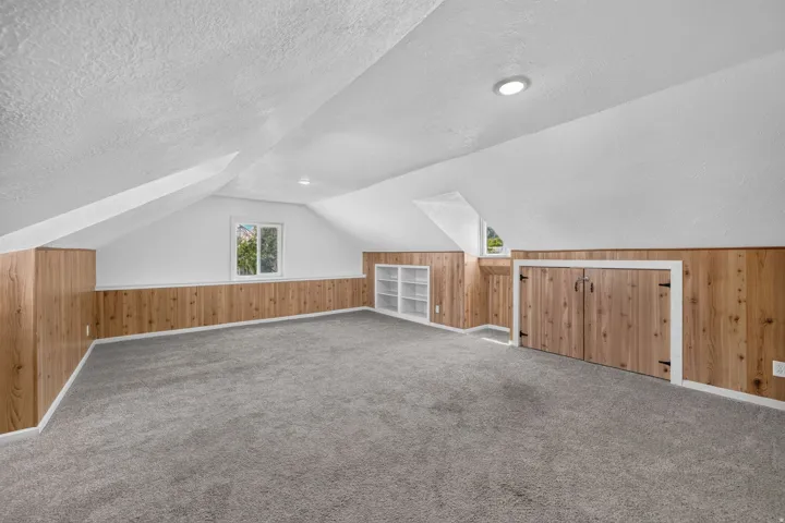 Spacious room featuring sloped ceilings, two windows, wood-panel accent walls, and soft grey carpeting