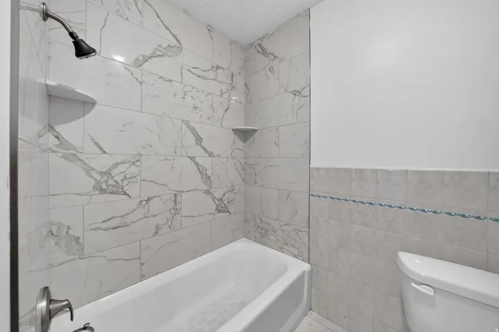 Bathtub and shower combination with floor-to-ceiling marbled tile surround, featuring a built-in shower niche and corner shelf
