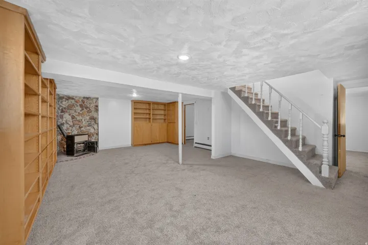 Spacious carpeted room featuring a stone accent wall, built-in shelving, a built-in wood stove, a staircase with white spindles, and recessed lighting