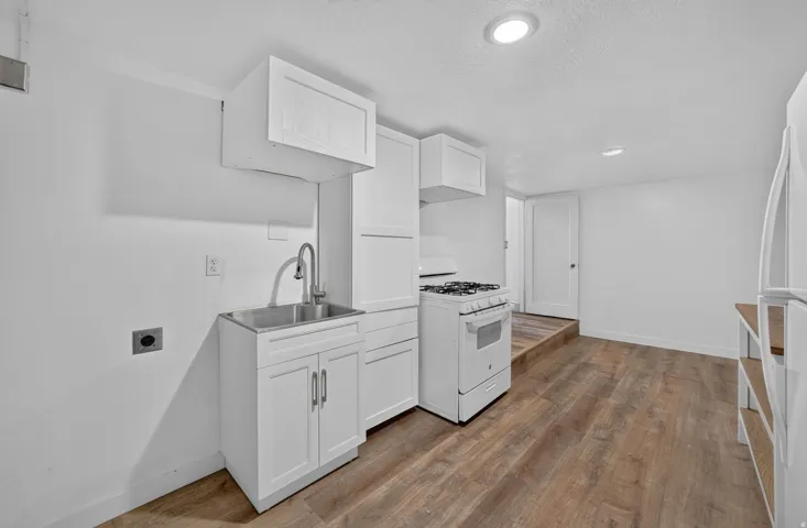 Compact kitchen area featuring wood-finish flooring, white cabinetry, a stainless steel utility sink with a high-arc faucet, a white gas range, and a white refrigerator