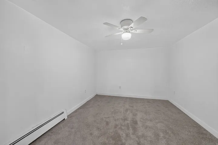Neutral tone interior featuring a ceiling fan with light fixture, baseboard heating, clean white walls, and gray carpet flooring