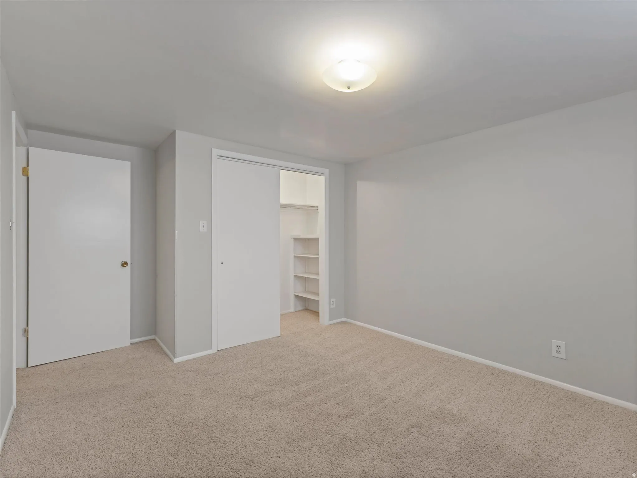 Neutral tone wall paint, light tan carpeting, flush-mount ceiling light fixture, and a built-in closet with shelving