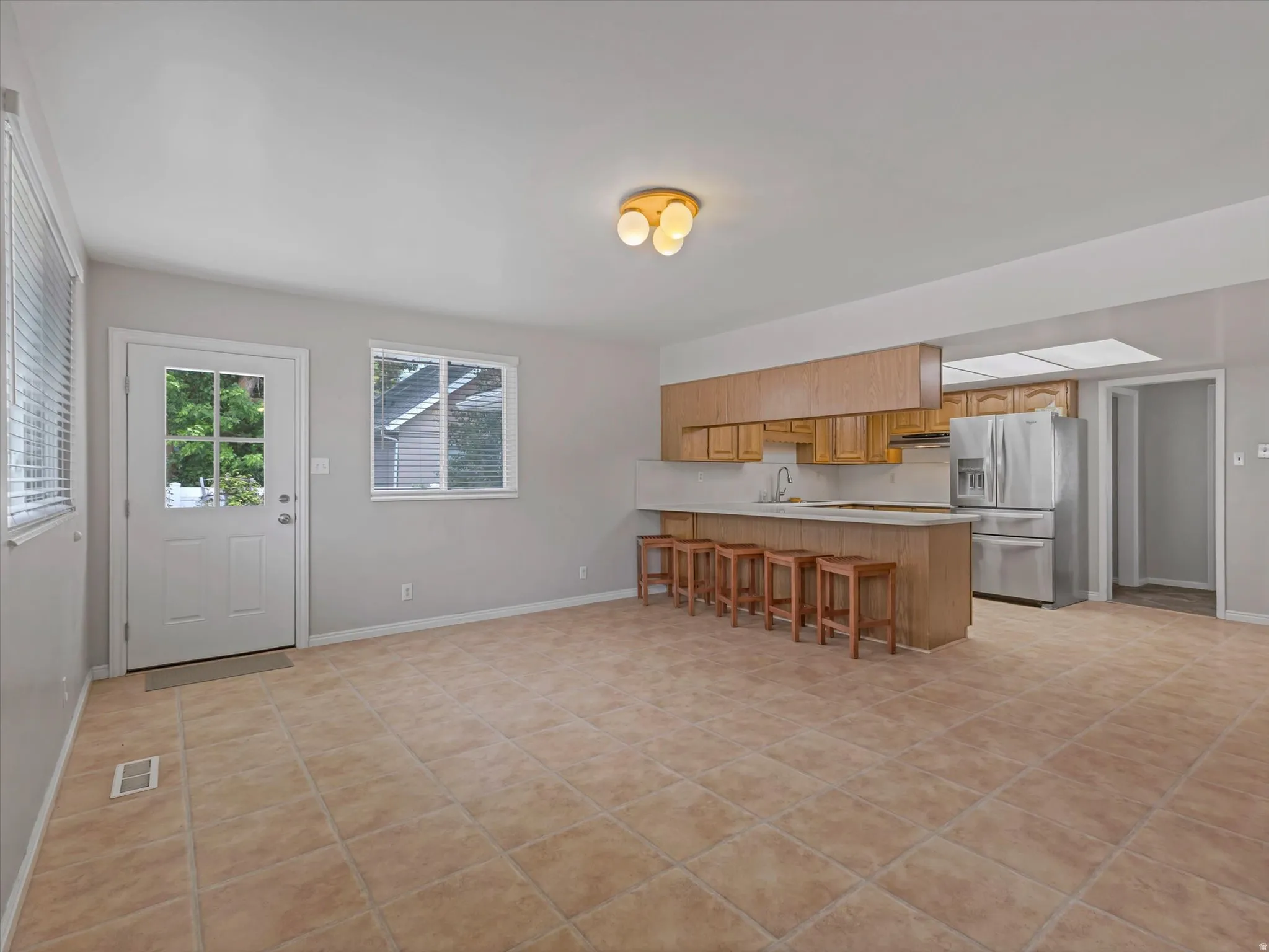 Open concept living space featuring light-toned tile flooring, a kitchen island with seating, natural wood-finish cabinetry, stainless steel appliances, and a single-light door