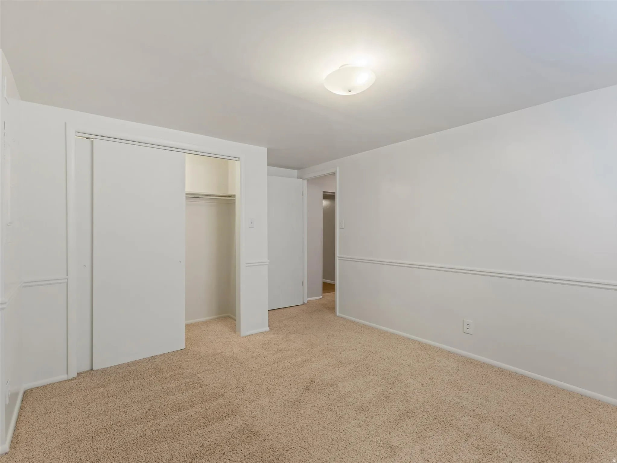 Spacious room featuring neutral carpeting, white walls with chair rail molding, a flush-mount ceiling light fixture, and a closet with sliding doors