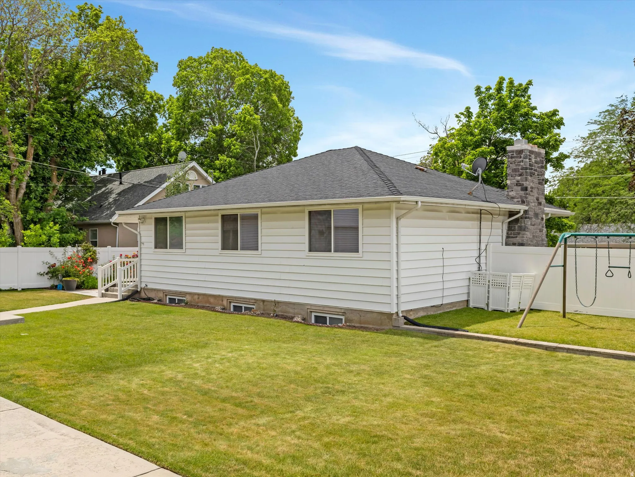 Expansive backyard featuring a manicured lawn, white vinyl fencing, a stone-clad chimney, and a small exterior stairwell leading to a back door