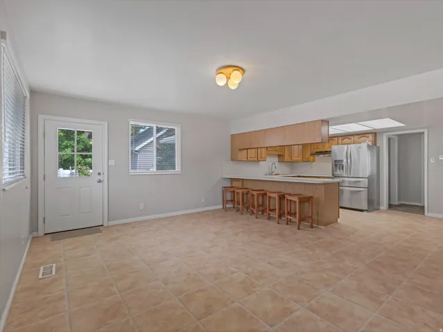 Open concept living space featuring light-toned tile flooring, a kitchen island with seating, natural wood-finish cabinetry, stainless steel appliances, and a single-light door