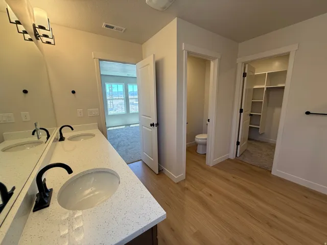 Dual vanity with white speckled countertop and undermount sinks, black hardware, wood-finish flooring, built-in closet shelving, and a separate water closet