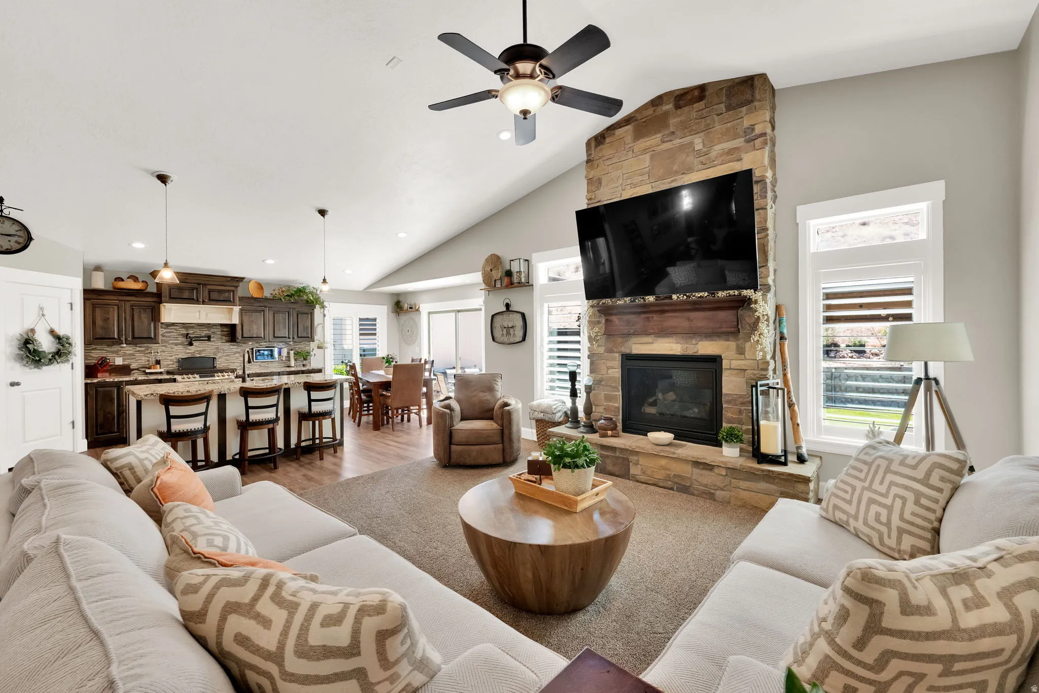 Vaulted ceiling living area featuring a stacked stone fireplace with a wood mantle, an integrated kitchen with a breakfast bar, recessed lighting, and wood-finish flooring