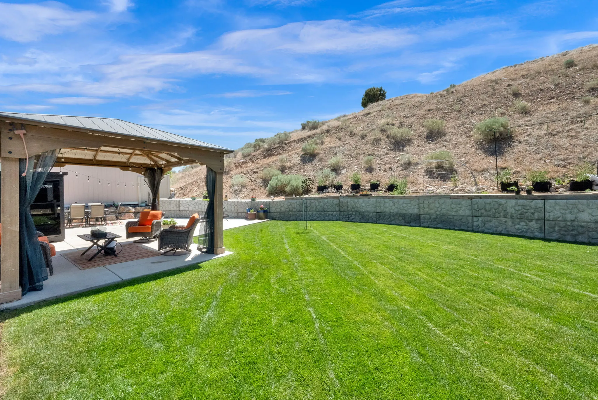 Expansive backyard featuring a large green lawn, a covered patio area with a wood-frame gazebo and privacy screens, a concrete patio surface, and a substantial stone retaining wall