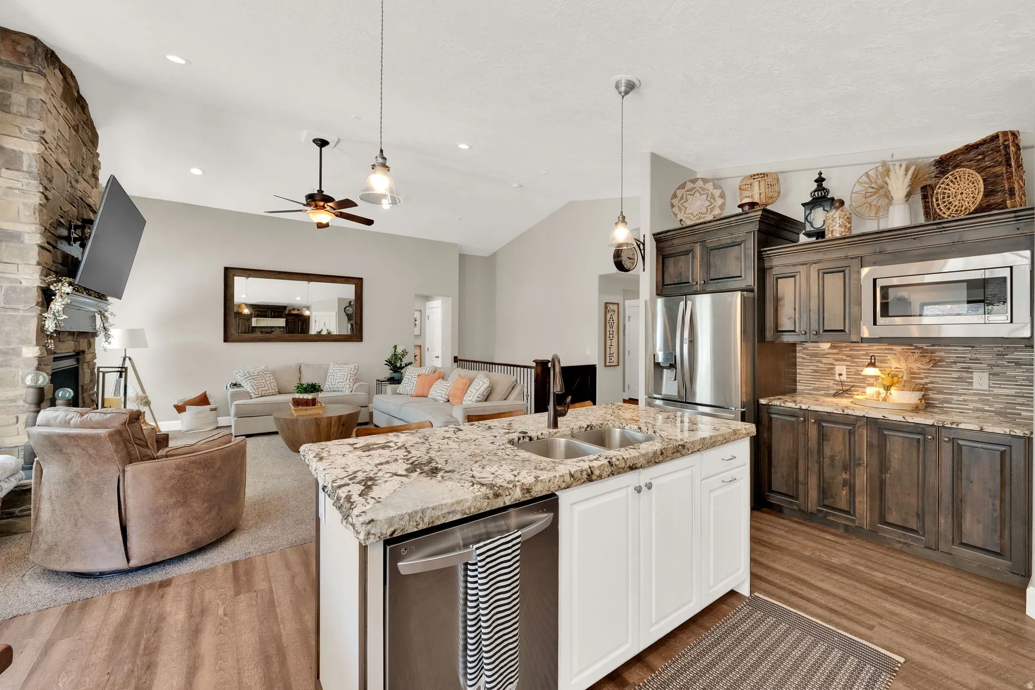 Open-concept kitchen and living area featuring granite countertops, stainless steel appliances, dark wood cabinetry, a stone fireplace, and vaulted ceilings