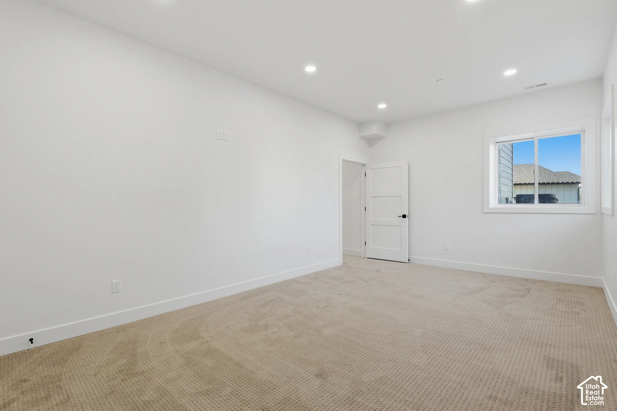 Spacious interior room featuring light-toned carpeting, white walls, recessed ceiling lighting, a contemporary white door, and a window providing natural light