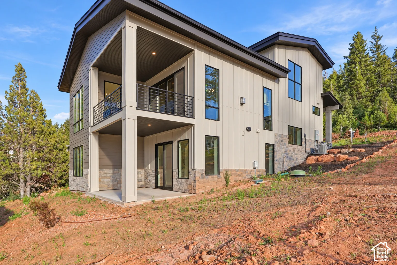 Modern architectural design featuring vertical siding, stone veneer accents, multiple large windows, a covered balcony with metal railings, and a covered patio