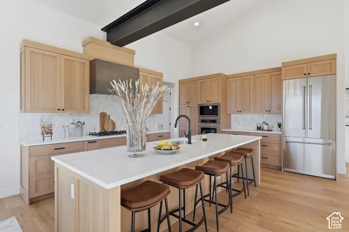 Gourmet kitchen featuring a large central island with seating, stainless steel appliances, light wood cabinetry, white countertops, and a striking exposed ceiling beam