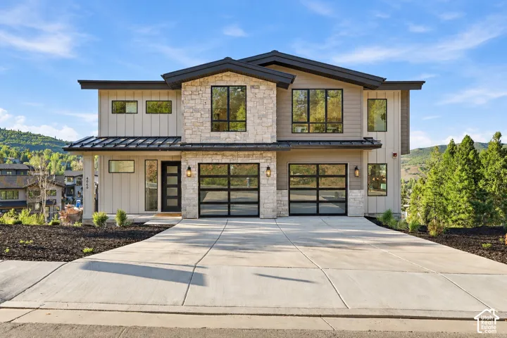 Contemporary facade featuring stone accents, vertical siding, multi-pane windows, a dark metal roof, and glass-paneled garage doors