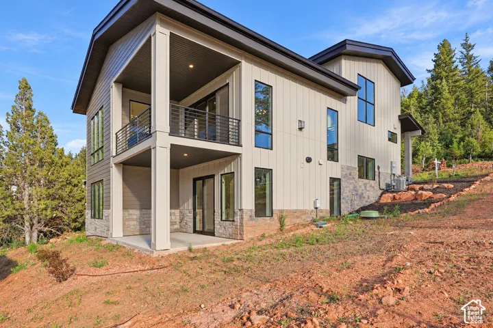 Modern architectural design featuring vertical siding, stone veneer accents, multiple large windows, a covered balcony with metal railings, and a covered patio