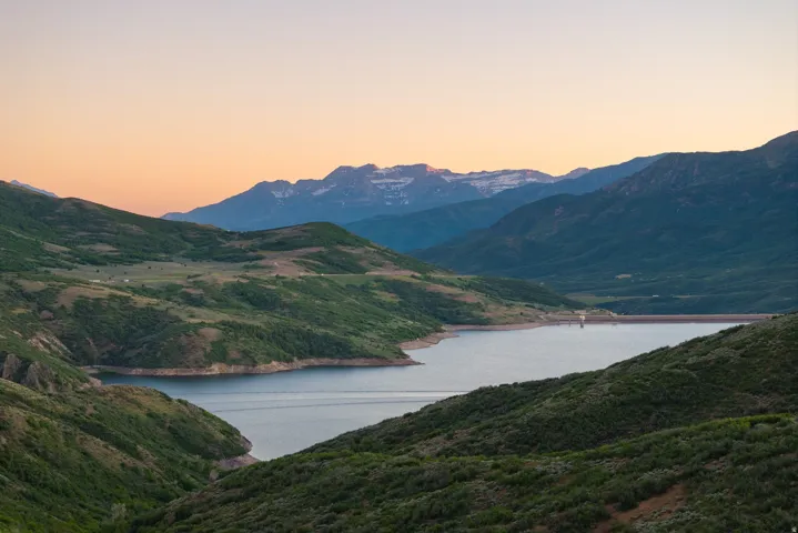 Expansive mountain landscape featuring a large body of water, rolling hillsides, distant snow-capped peaks, and a clear horizon