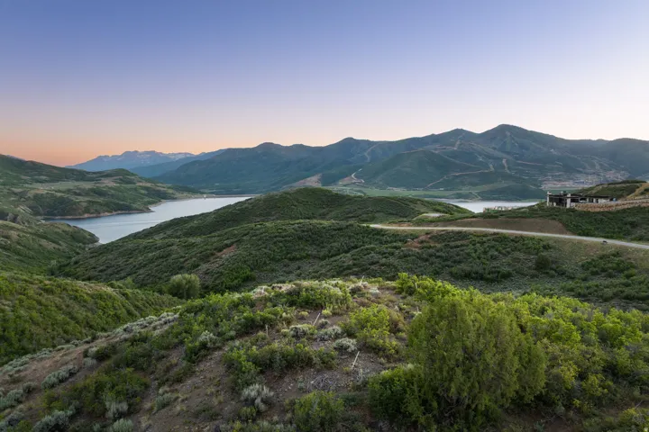 Expansive mountain terrain with a prominent body of water, distant mountain peaks, a winding road, and a contemporary structure with a flat roofline