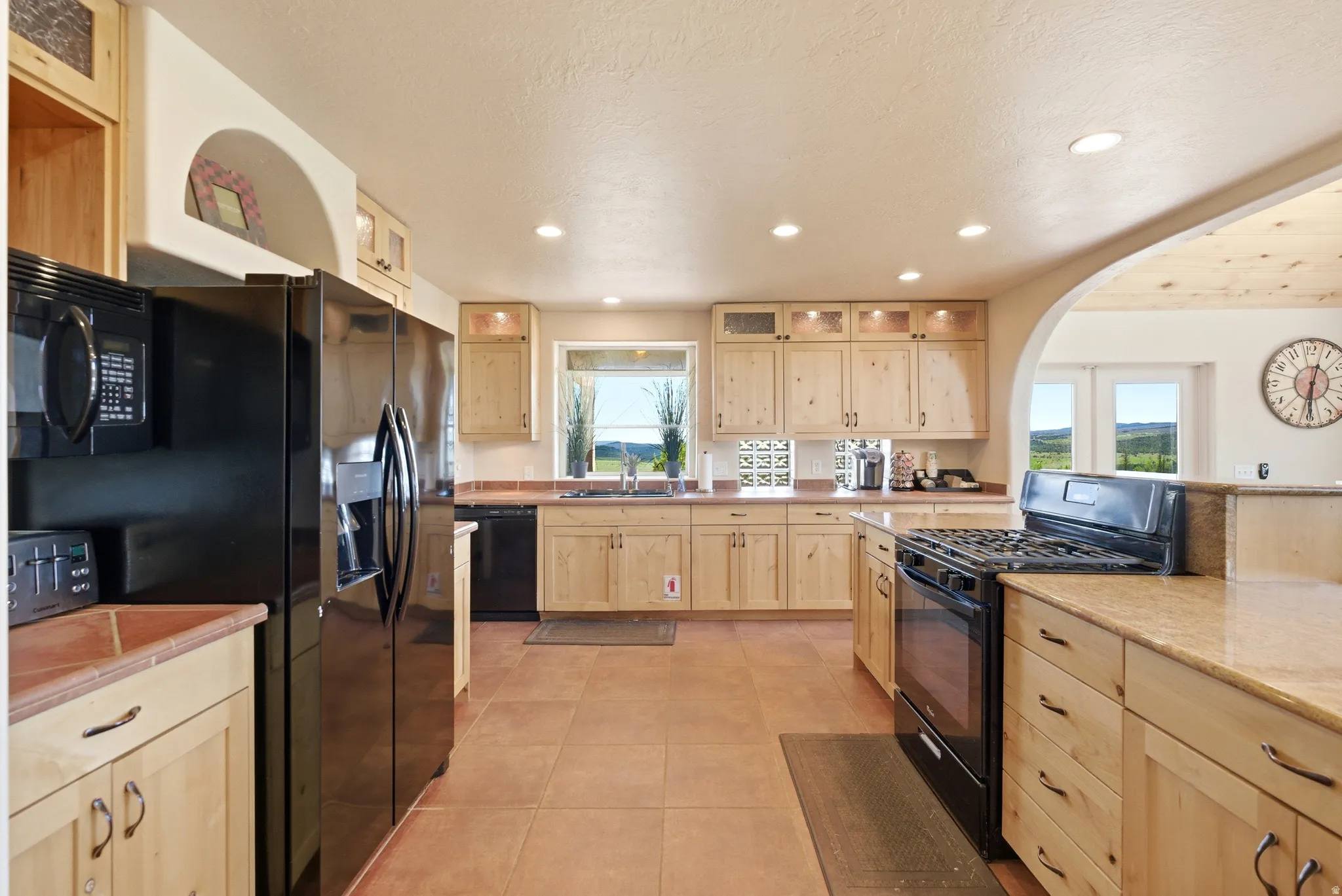 Expansive kitchen featuring wood cabinetry, stone countertops, integrated black appliances, arched architectural details, and tile flooring