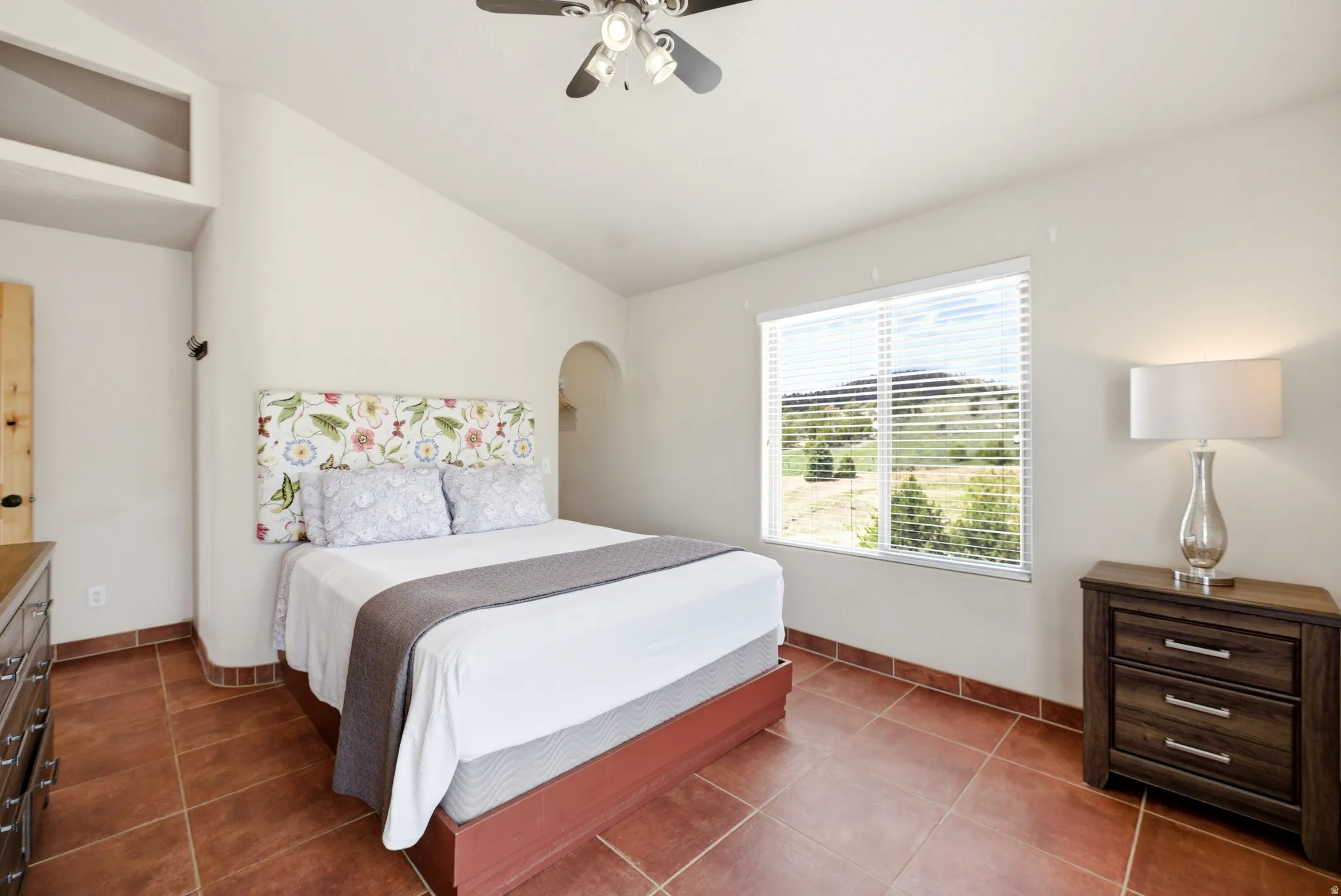 Spacious interior featuring a large window with horizontal blinds, terracotta tile flooring, vaulted ceilings, a ceiling fan, and an arched wall niche