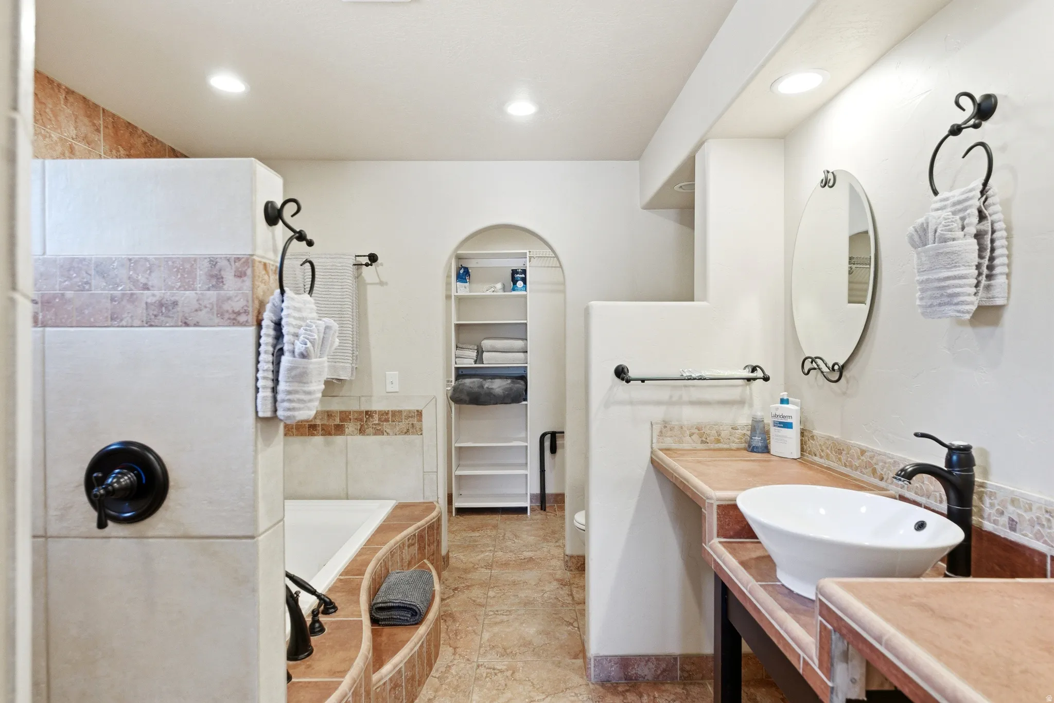Bathroom featuring a built-in soaking tub, tiled shower surround with accent band, vessel sink vanity with tiled countertop, arched alcove shelving, and recessed lighting