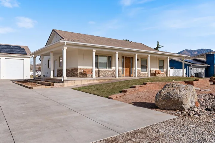 Ranch-style residence featuring a wide concrete driveway, an attached garage with solar panels, a covered front porch with pillars, and stone veneer accents