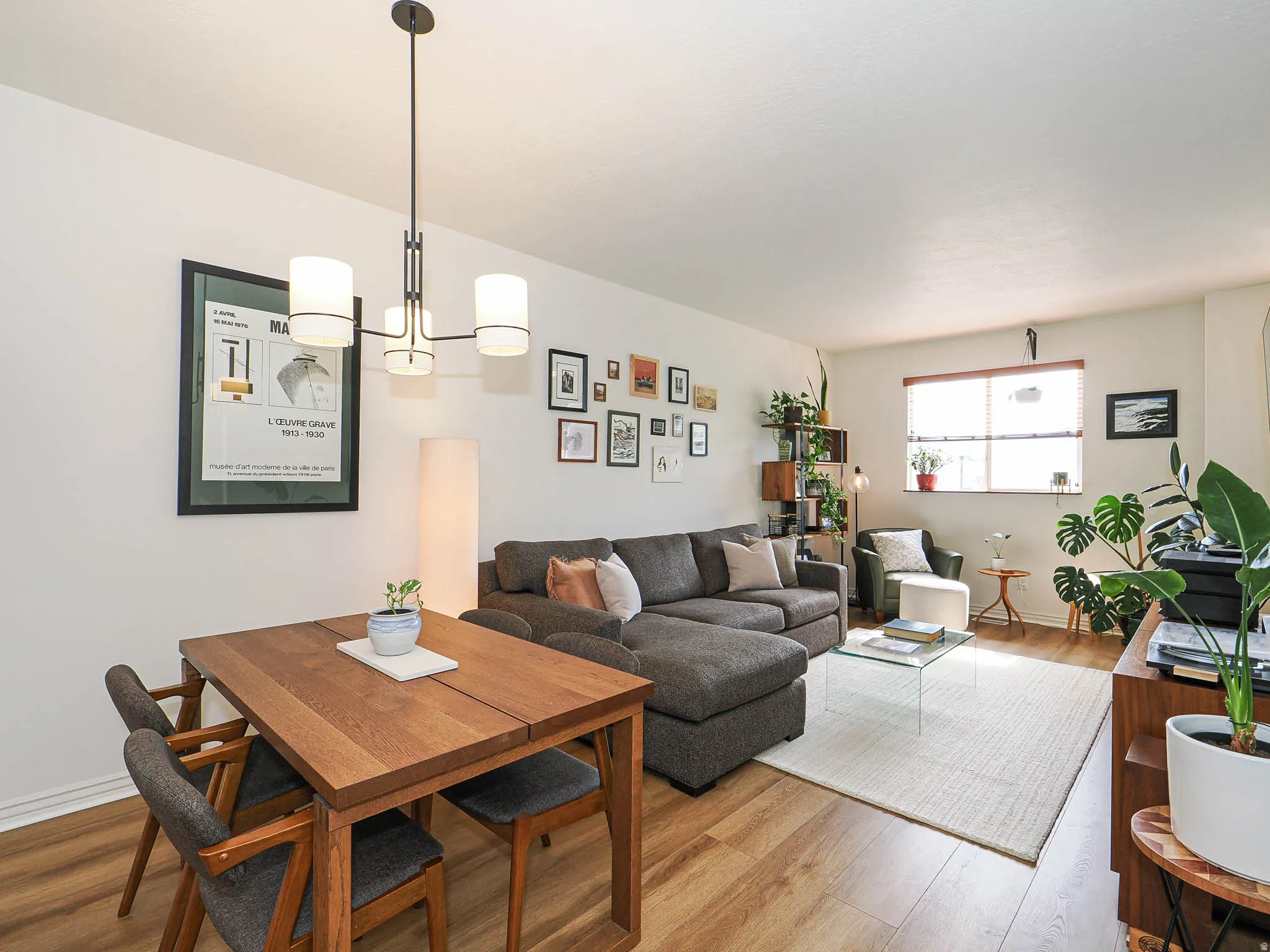 Open-concept living area featuring wood-finish flooring, a contemporary chandelier, and a large window with blinds